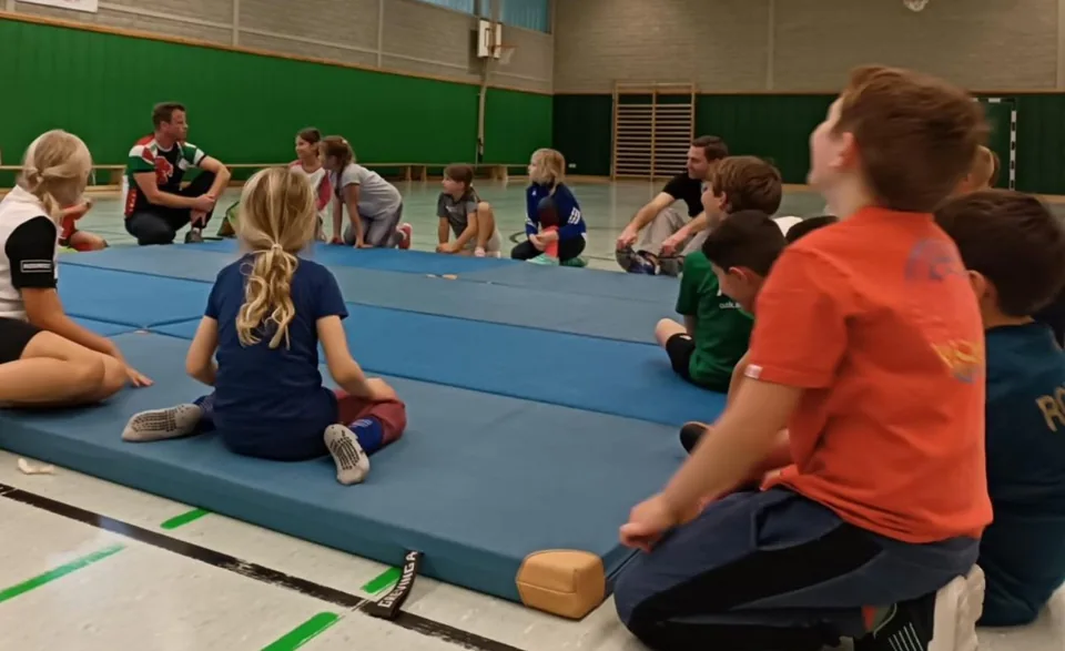 Oliver sitzt mit einer Kindergruppe und einem weiteren Erwachsenen im Kreis auf blauen Matten in einer Sporthalle.