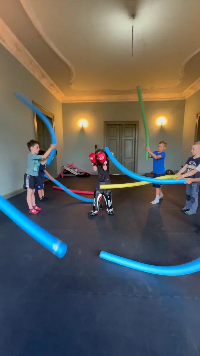 Kinder bewegen sich bei einer Übung mit bunten Schaumstoffstangen im Trainingsraum.